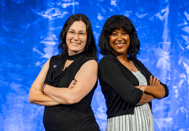 Bobbi Kay and Mahoganey Jones stand back to back smiling at the camera against a blue wall. 