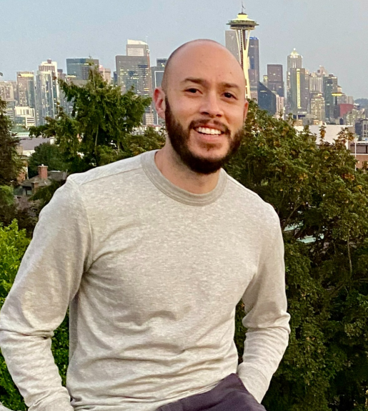 Daniel Acosta pictured in front of a city skyline with a long-sleeve light shirt, smiling with a short beard.