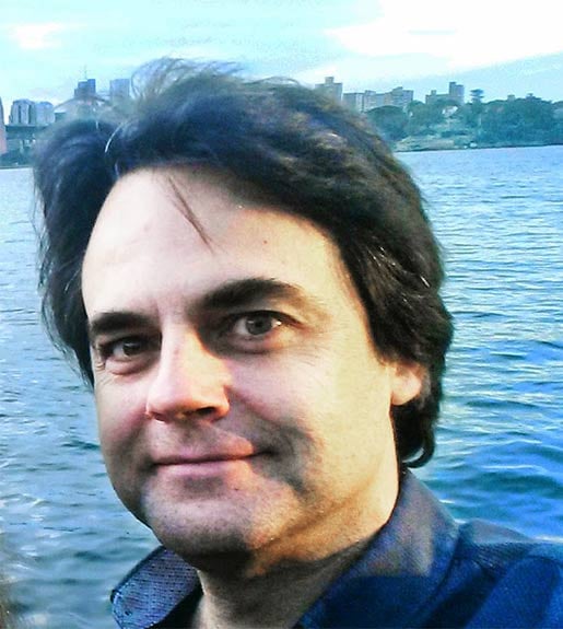 A person with dark hair, wearing a blue shirt, seen from the neck up. A body of water in the background.