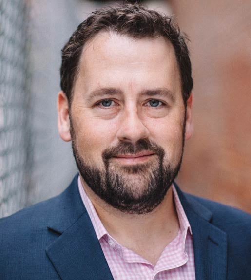 Headshot of a person with short brown hair, a trim beard and mustache, wearing a navy blazer and pink button-up shirt.