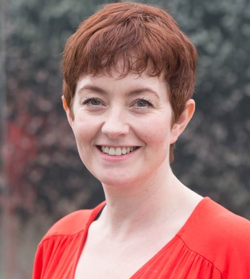 Photo of a woman from the shoulders up. She has short, dark red hair and is facing forward and smiling.