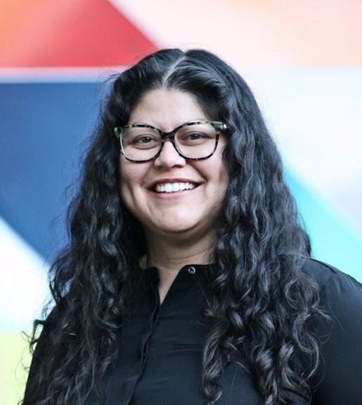 A person smiling with long, dark, curly hair and dark framed glasses, and wearing a dark shirt in front of a colorful background.