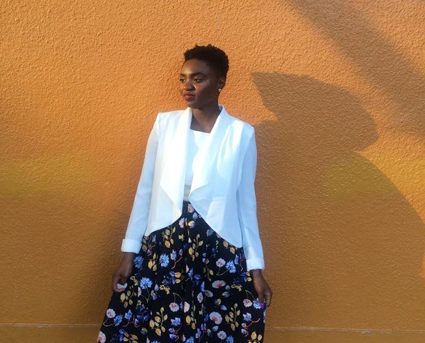 Tia Bangura, a Black woman with short black hair, wearing a white top and floral print black skirt, standing in front of an orange wall.