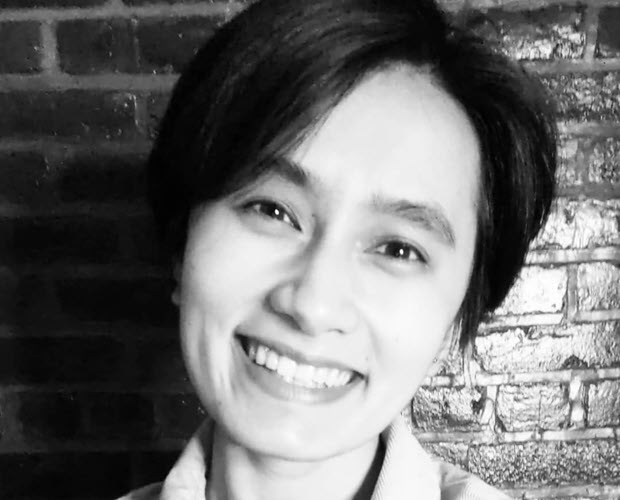 Black and white photo of Kelley Ho, a Vietnamese American woman with short dark hair, in front of a brick wall.