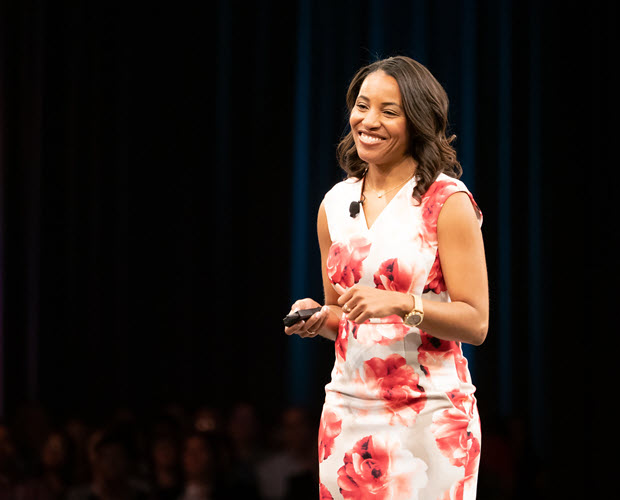 Nicole Smith, a black woman with long dark hair wearing a dress with a large floral print, seen speaking on stage at a conference.