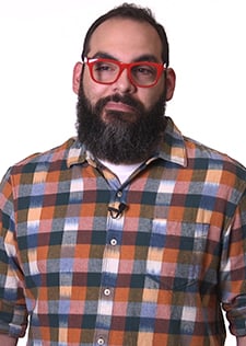A bearded man with brown hair and red glasses wearing a plaid shirt