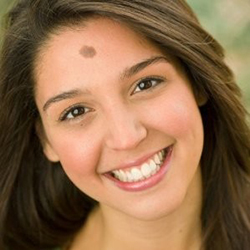 Anne Mortomore, a young woman with long, brown hair and a birthmark on her forehead, smiles directly at the camera