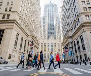 A group of people walk through an intersection. They are surrounded by tall buildings.