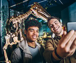 Two young men taking a selfie in front of a large skeletal dinosaur fossil at the National Museum of Wales.