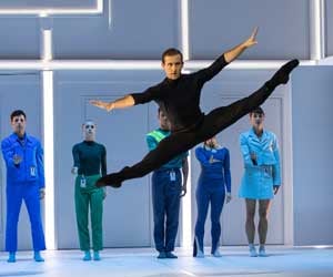 A male dancer wearing black leaps across a white stage. 