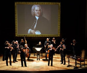 Several musicians, most of them standing, perform under a large screen showing an image of J.S. Bach