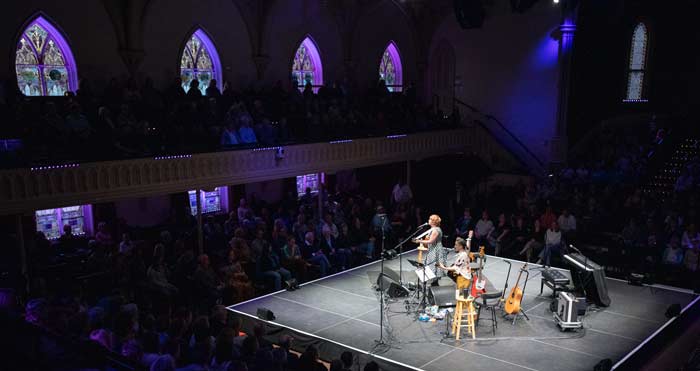Photo of an ensemble playing on a square stage, surrounded by seated audiences on all sides.