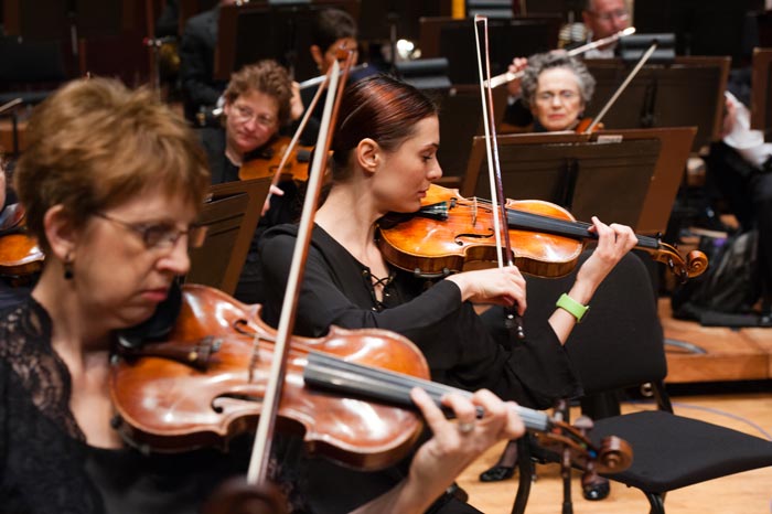Women playing the violin, with other orchestra members (including a flutist) behind them