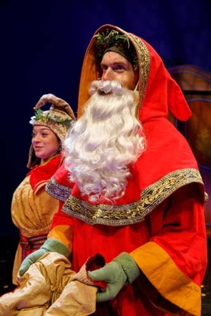 An actor in a red hooded costume and long white fake beard, facing left and speaking.