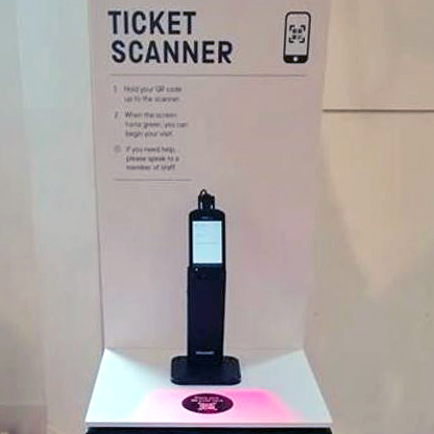 Photo of a small black device on a stand, with a sign behind it reading Ticket Scanner and showing a mobile phone with a square barcode on it.