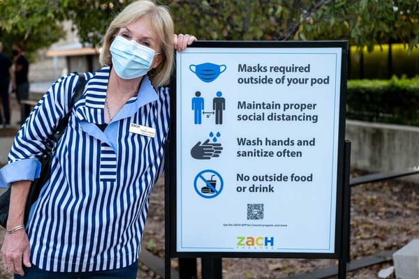 A woman wearing a mask standing next to a sign listing four safety protocols, including mask requirements, physical distancing, and washing & sanitizing hands frequently.