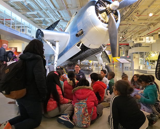 A group of school children listen to a tour guide in front of a large airplane at the Intrepid Sea, Air and Space Museum.