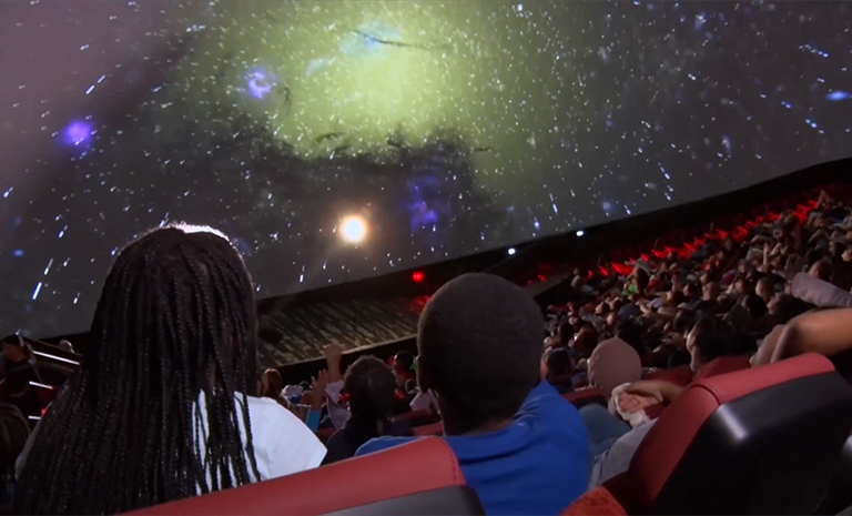 A mother and son seen from the back react to images of the solar system on the big screen of the Liberty Science Center planetarium.