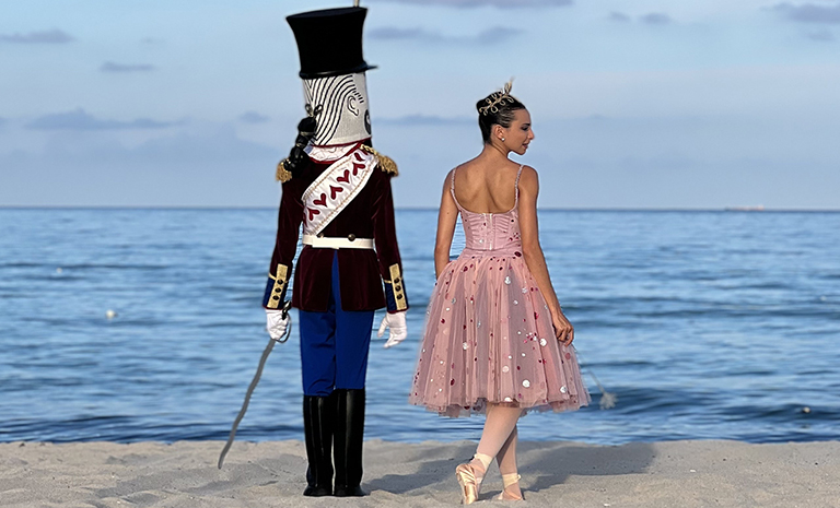 Two dancers, one costumed as the Nutcracker and the other in pink as the Sugar Plum Fairy, stand at the edge of a sandy beach gazing out into the ocean.