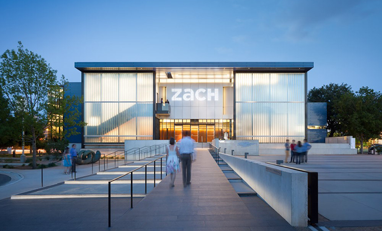 A man and woman walk toward the entrance of ZACH Theatre, a building with a large glass facade, at twilight.
