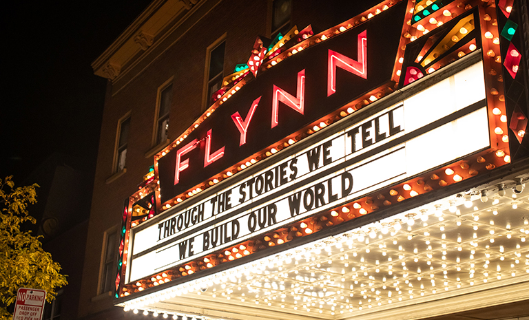 Marquee sign for the Flynn Center for the Performing Arts, lit at night and displaying the words 'Through the stories we tell, we build our world.'