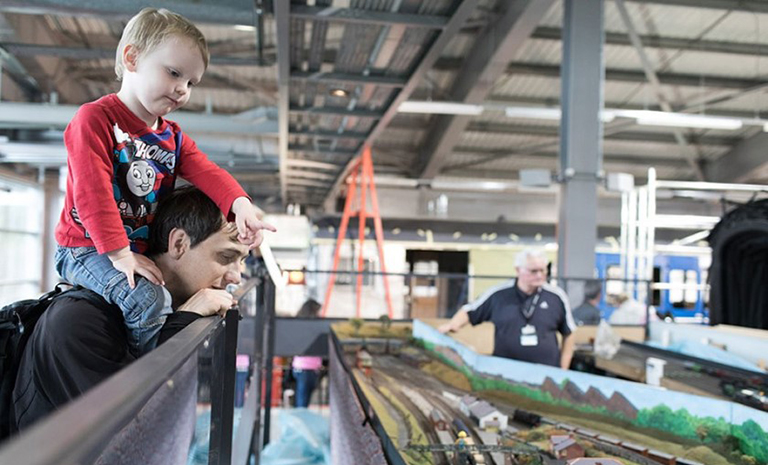 A father with his son on top of his shoulders, pointing, look at a model train set at Locomotion museum in the UK.