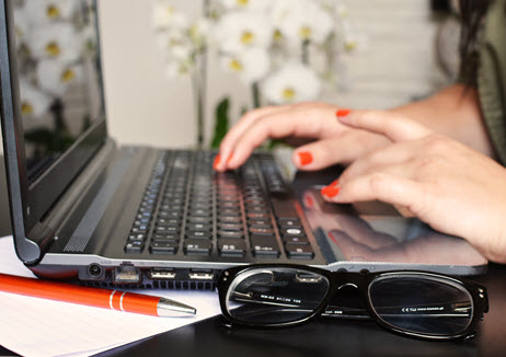 A close-up of two hands, wearing red fingernail polish, typing on a laptop. In the foreground, a pair of eyeglasses on the table.