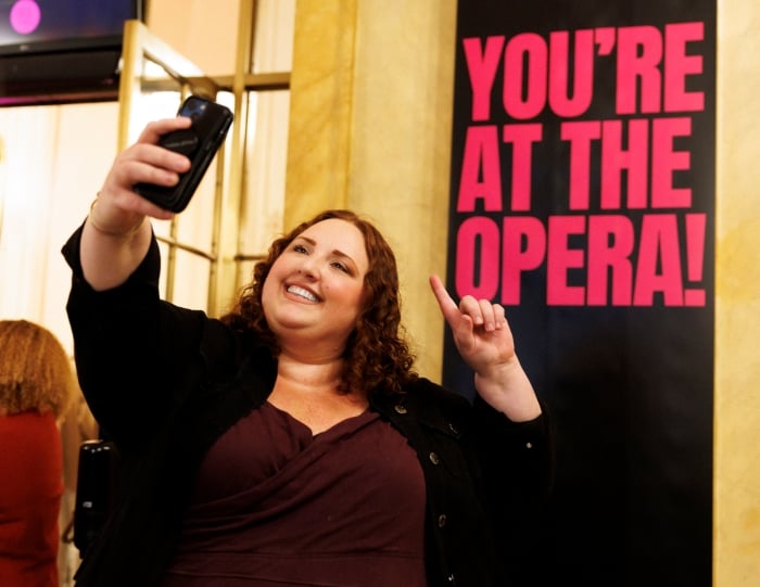 A guest happily poses for a selfie in front of a sign that reads You're at the opera! in capitalized, red letters at Opera Philadelphia. 