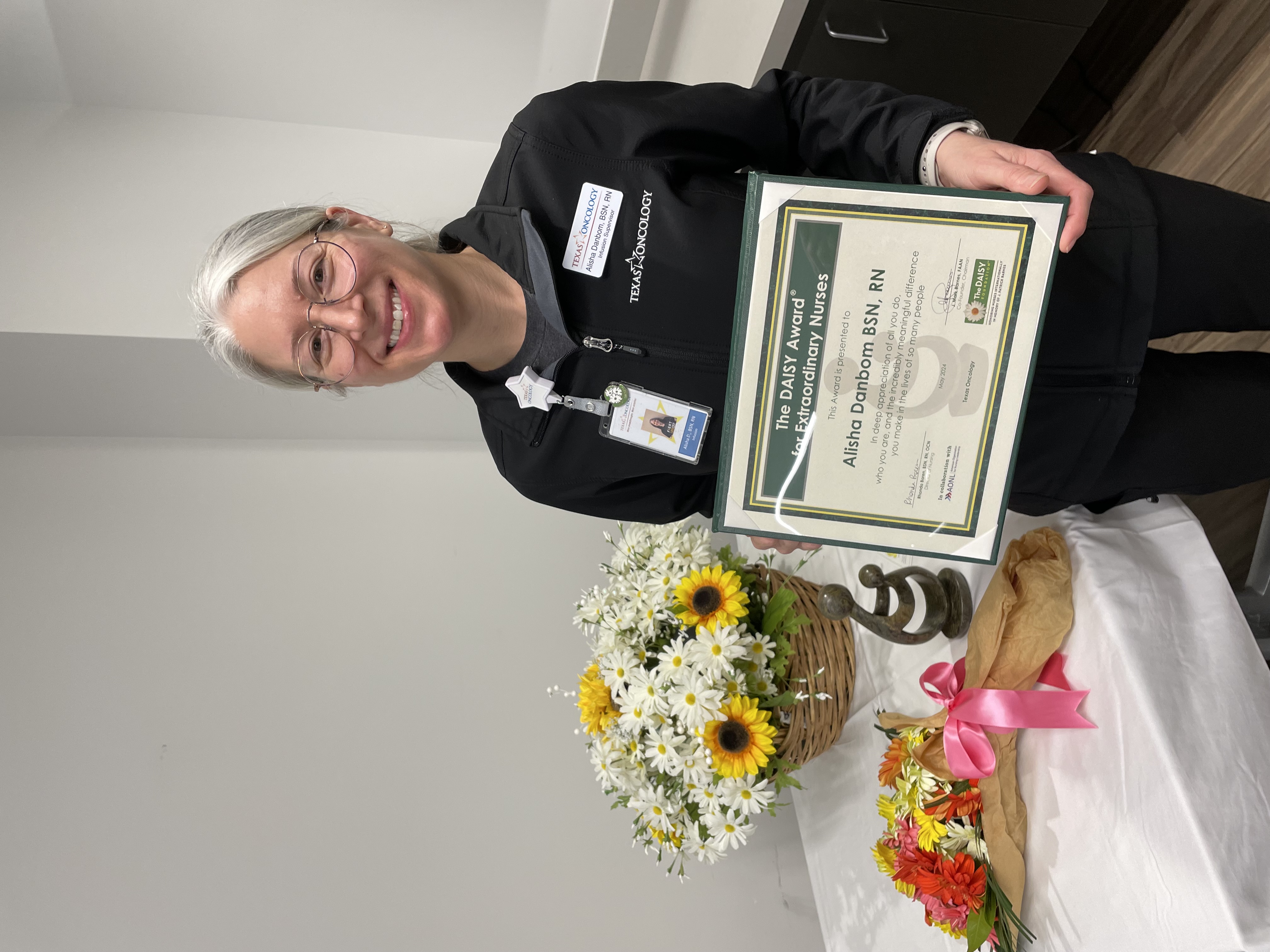 Alisha Danbom, BSN, RN, smiling in glasses and a black Texas Oncology jacket, holding her framed DAISY Award certificate for Extraordinary Nurses beside a sunflower and daisy arrangement and the DAISY sculpture.