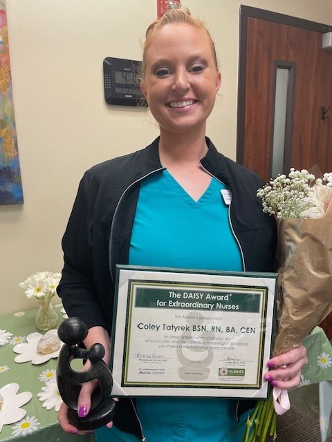 Coley Tatyrek, BSN, RN, BA, CEN, smiling in a black jacket over teal scrubs, holding the DAISY sculpture and her award certificate for Extraordinary Nurses beside a bouquet of flowers on a daisy-decorated table.