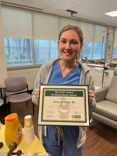 Emily Ford, BSN, RN, smiling in a blue Texas Oncology scrub jacket, holding her DAISY Award certificate for Extraordinary Nurses in an infusion room.