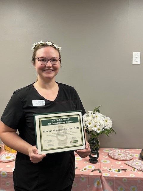 Hannah Kingcade, BSN, RN, OCN, smiling in glasses and a daisy flower crown, holding her DAISY Award certificate for Extraordinary Nurses at a table decorated with a pink daisy-print tablecloth.