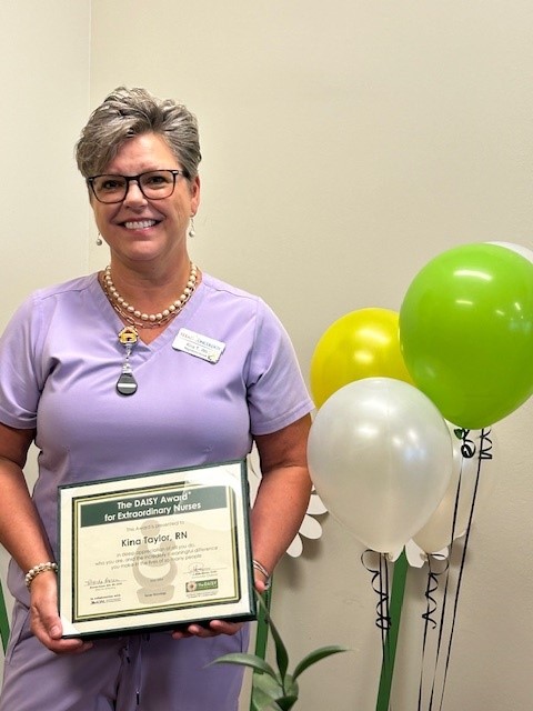 Kina Taylor, RN, smiling in glasses, a lavender scrub top, and a pearl necklace, holding her DAISY Award certificate for Extraordinary Nurses in front of green, yellow, and white balloons.