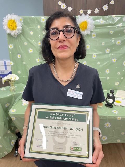 Ladan Ghadiri, BSN, RN, OCN, smiling in glasses and navy Texas Oncology scrubs, holding her DAISY Award certificate for Extraordinary Nurses in front of a green daisy-print tablecloth and paper flower decorations.