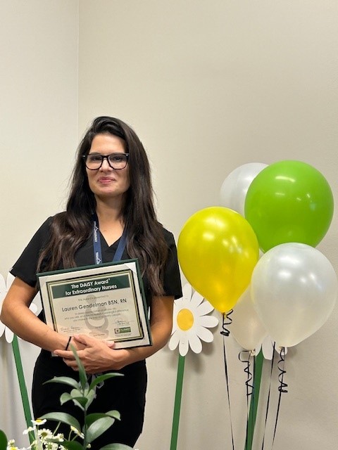Lauren Gendelman, BSN, RN, smiling in glasses and a black top, holding her DAISY Award certificate for Extraordinary Nurses in front of green, yellow, and white balloons and paper daisy cutouts.