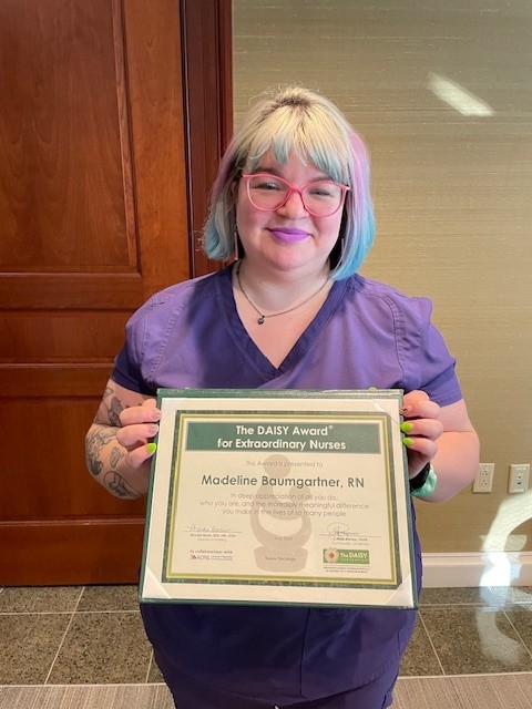 Madeline Baumgartner, RN, smiling in glasses, purple scrubs, and pink and blue hair, holding her DAISY Award certificate for Extraordinary Nurses in a Texas Oncology hallway