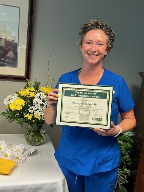 Meagan Jones, RN, smiling in blue scrubs, holding her DAISY Award certificate for Extraordinary Nurses beside a vase of yellow and white daisies.