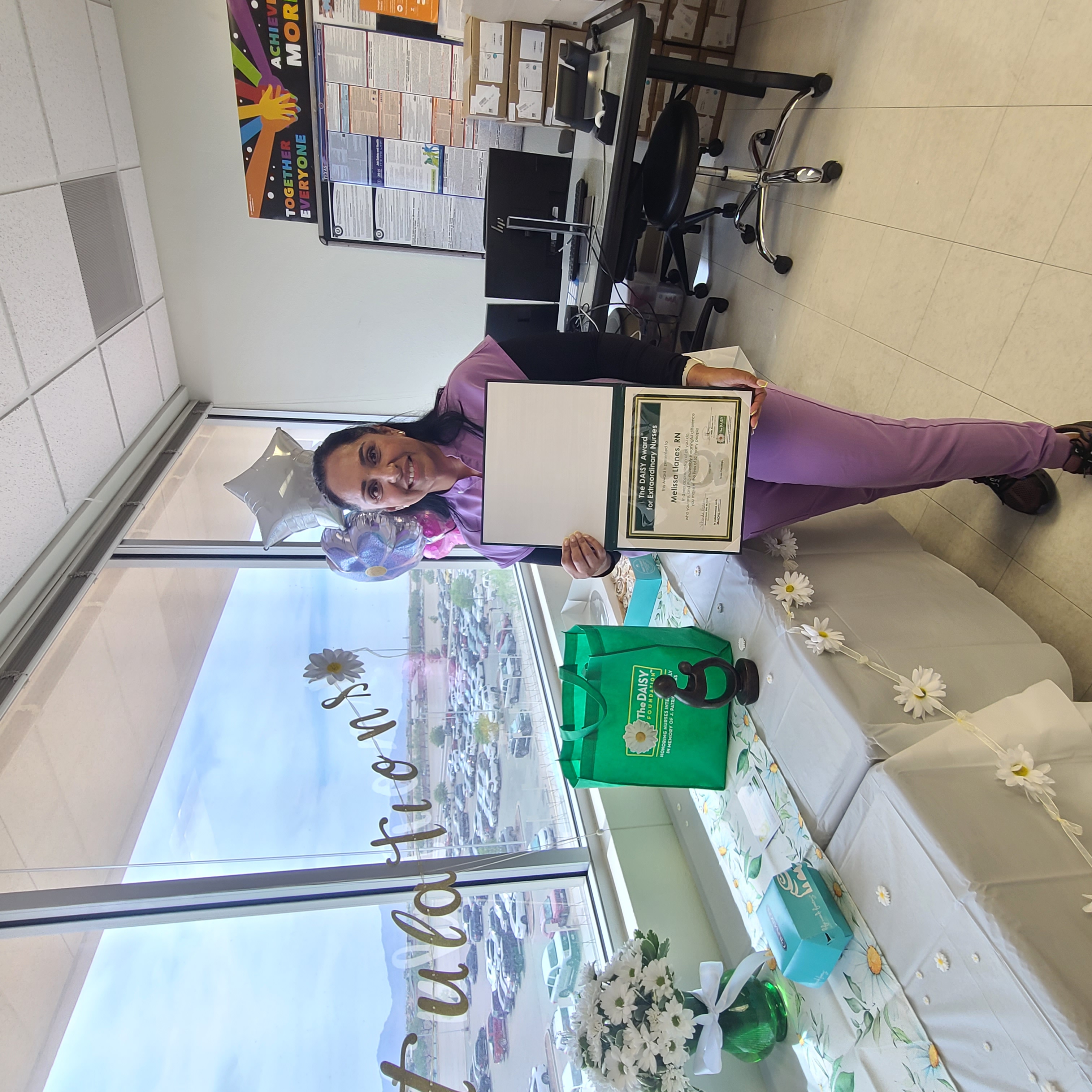 Melissa Llanes, RN, smiling in a lavender scrub top, holding her DAISY Award certificate and the DAISY sculpture in a Texas Oncology office decorated with a Congratulations window banner and silver star balloons. 
