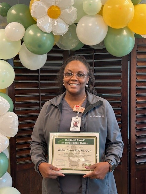 Melissa Sykes, BSN, RN, OCN, smiling in glasses and a gray Texas Oncology jacket, holding her DAISY Award certificate for Extraordinary Nurses in front of a green, yellow, and white balloon flower arch.
