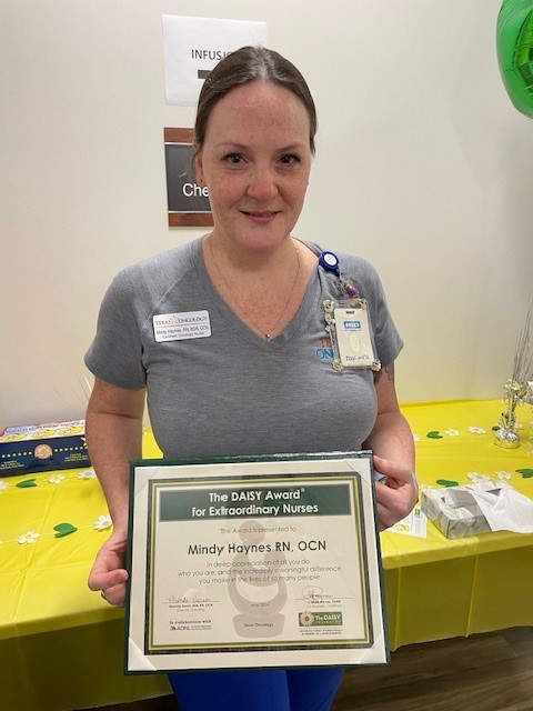 Mindy Haynes, RN, OCN, smiling in a gray Texas Oncology top, holding her DAISY Award certificate for Extraordinary Nurses in front of a table decorated with daisies near the infusion area.