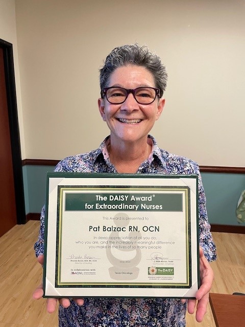 Pat Balzac, RN, OCN, smiling in glasses and a floral shirt, holding her framed DAISY Award for Extraordinary Nurses at Texas Oncology.