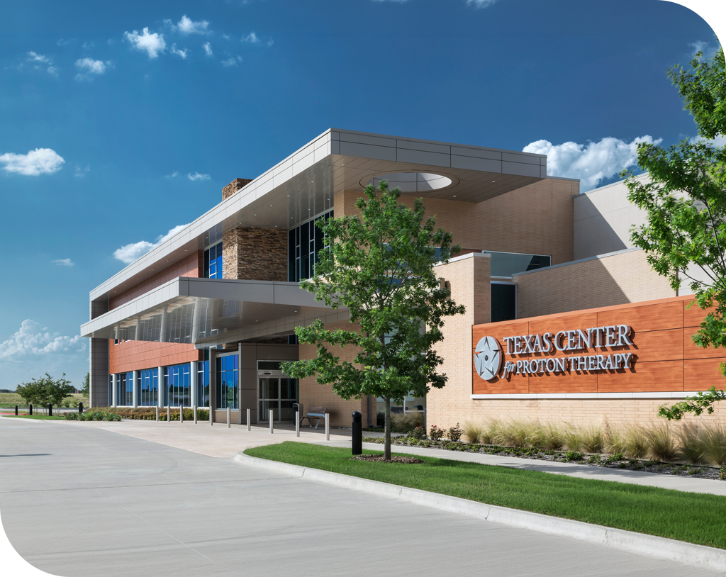 Exterior of Texas Oncology’s Texas Center for Proton Therapy in Irving, Texas, a modern two-story building with a covered entrance, stone and copper-toned accents, and a landscaped front lawn under a bright blue sky.