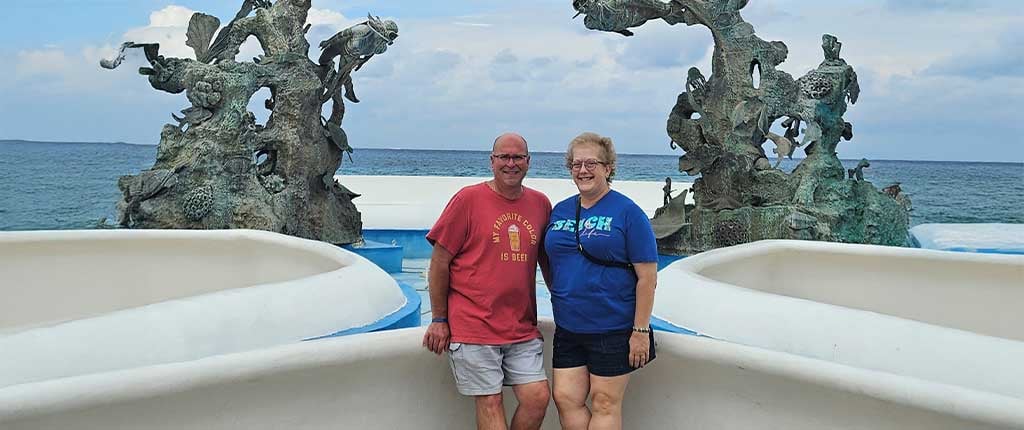 Debbie Goforth, a metastatic breast cancer survivor, stands smiling with her husband near an oceanfront sculpture, with the sea visible in the background.