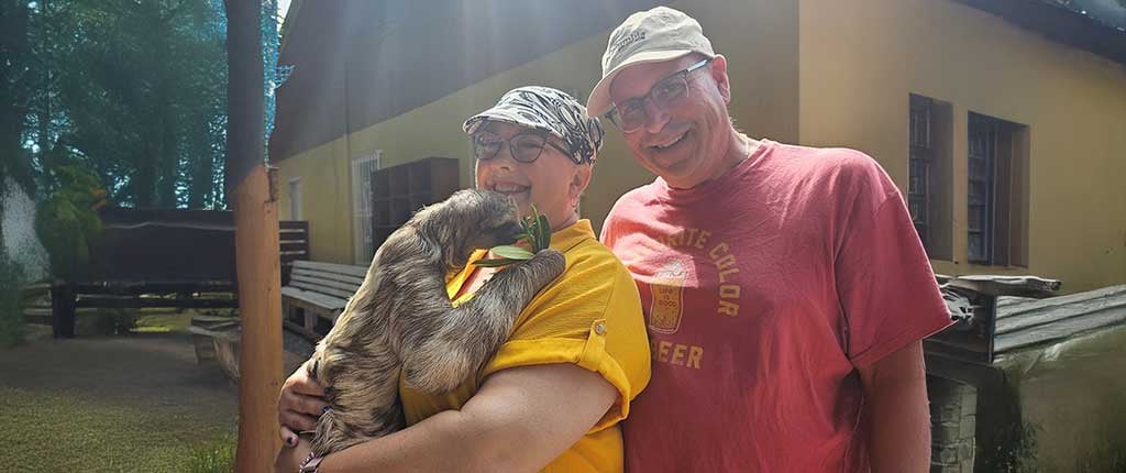 Debbie Goforth, a metastatic breast cancer survivor, smiles while holding a sloth, standing beside her husband outdoors in a sunny setting.