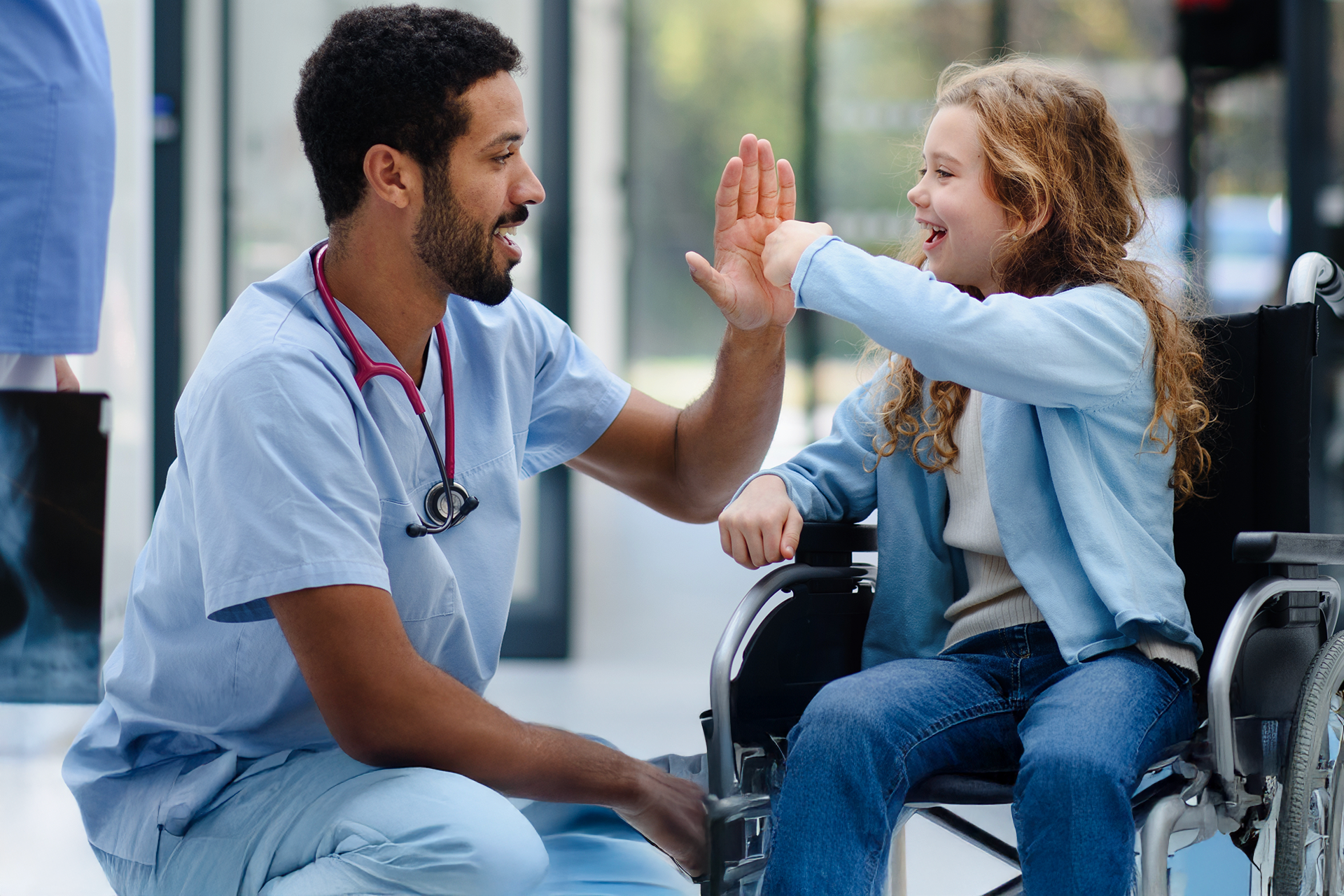 Child in a wheelchair high-fiving a smiling doctor in a hospital hallway.