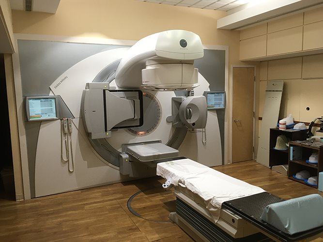 Modern radiation therapy treatment vault featuring a Varian linear accelerator with patient treatment couch, illuminated sky-scene ceiling panels for patient comfort, and wood-look flooring