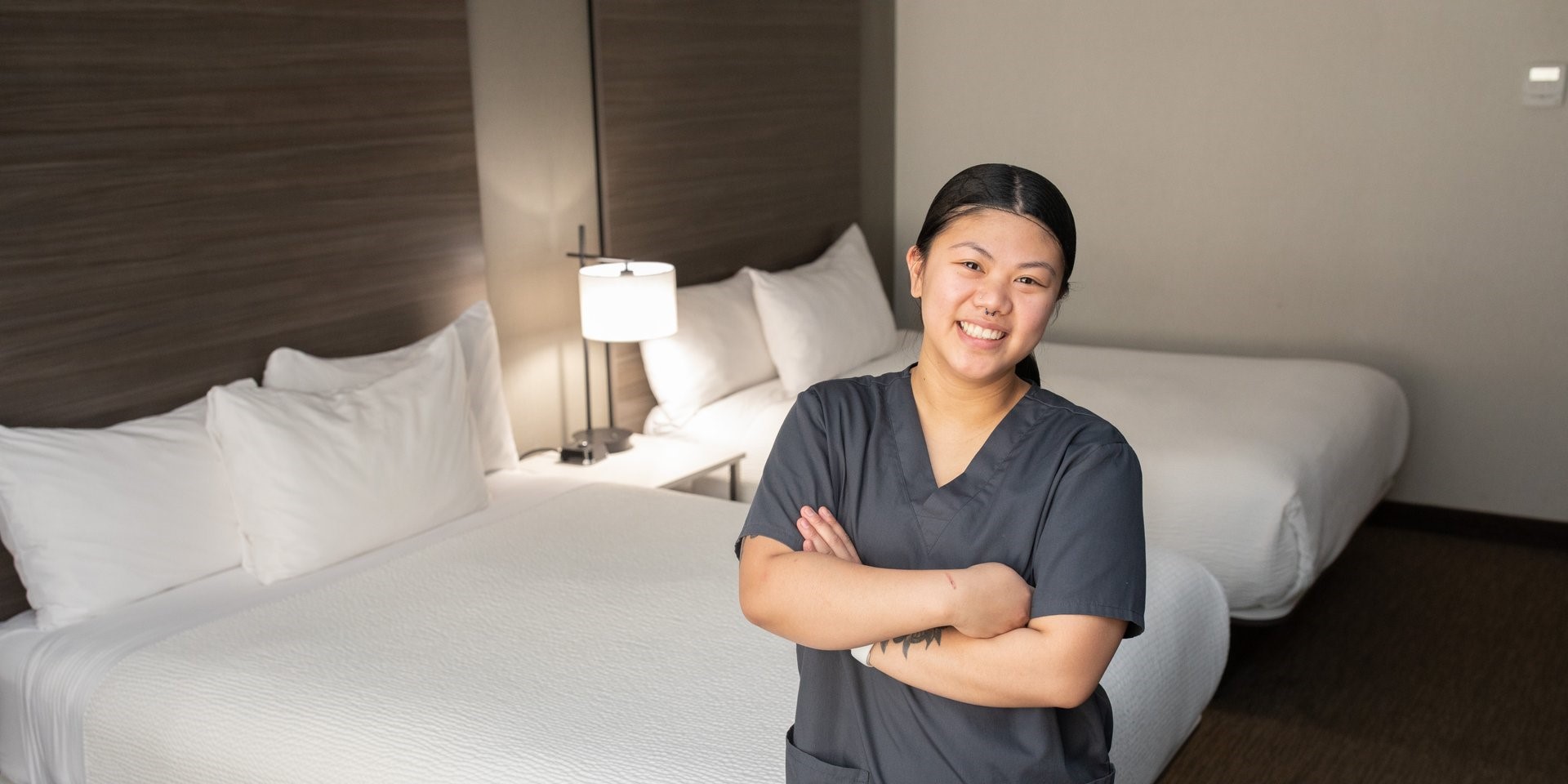 Woman in hotel uniform standing with arms folded and a smile in a neatly made up hotel room