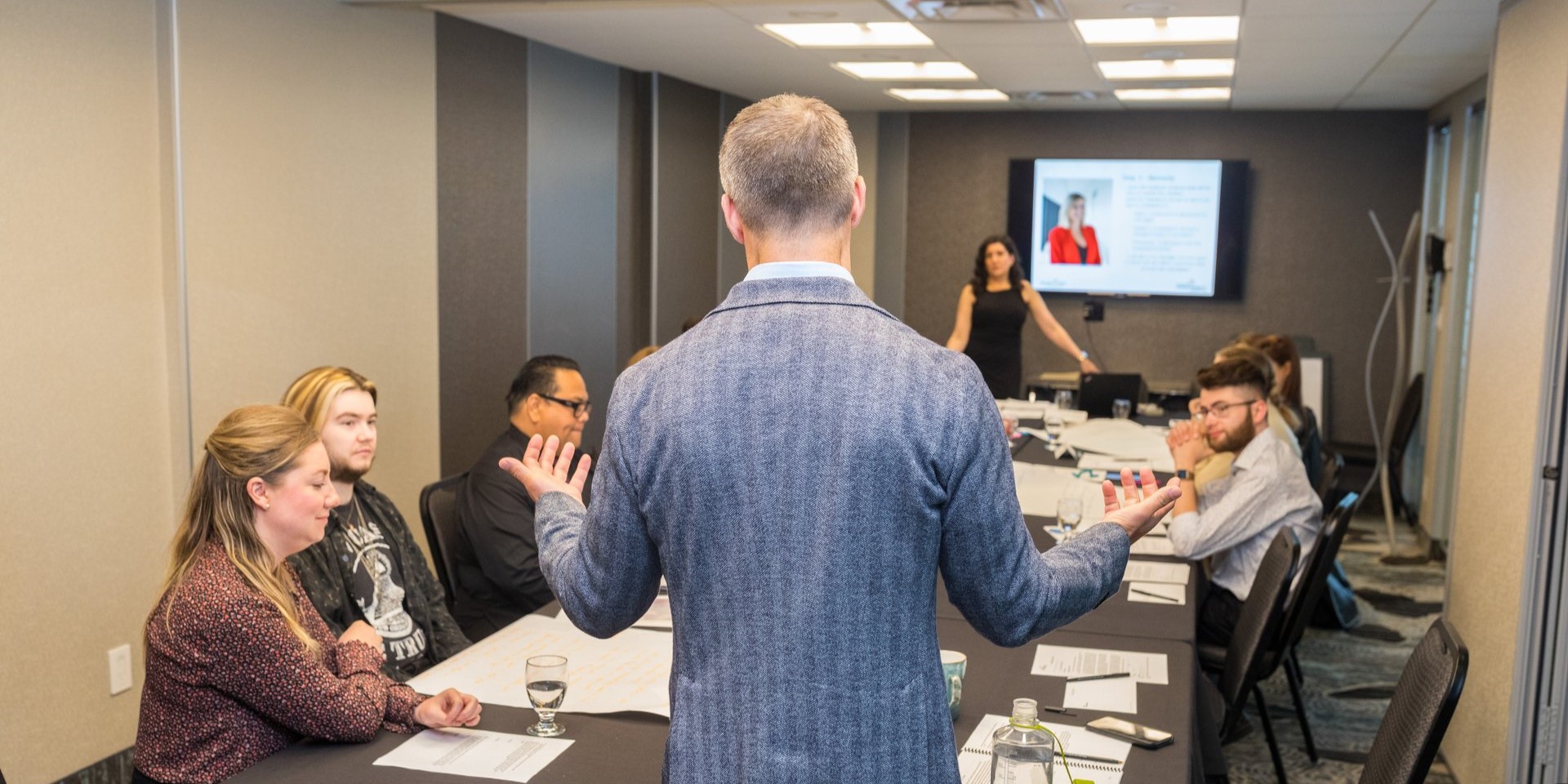 Tall man with short greying hair and wearing a blue herringbone suit jacket gestures with his palms raised toward a boardroom table that has a group of people seated around it, some of whom are looking at the man and others are looking at training materials on the table, while a classroom trainer who is a woman with dark hair wearing a dark dress at the opposite end of the table looks toward the camera and a television screen behind that is showing a slide with a course lesson