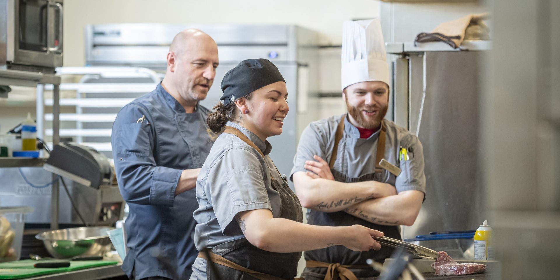 A man in executive chef uniform and a man in a cook uniform watch as another cook uses tongs to turn a steak on a hot grill