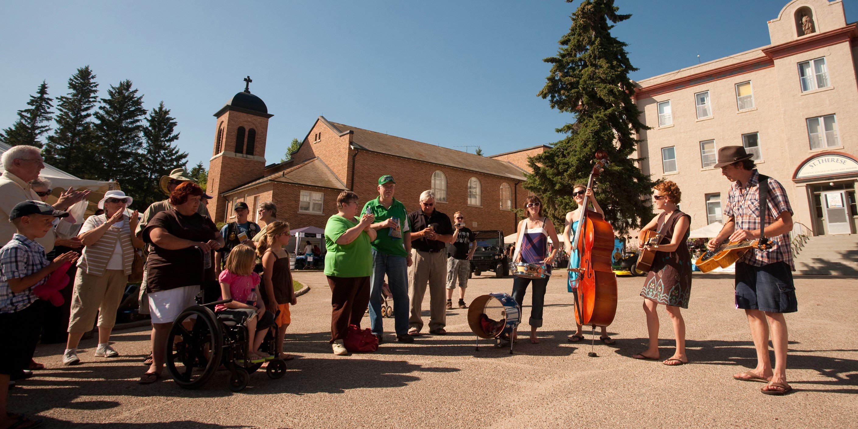 group of people of various ages standing and watching a group of four musicians perform in front of an old building under a bright sunny sky